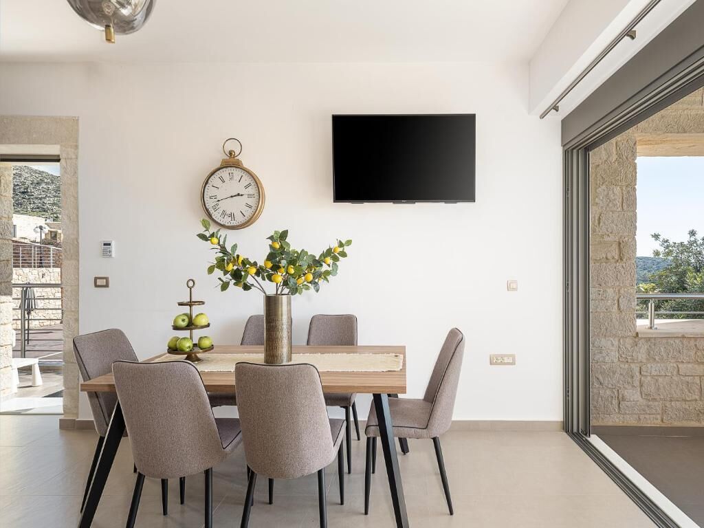 Un comedor moderno con una mesa de madera, sillas grises, un reloj montado en la pared y un televisor de pantalla plana.