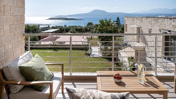A balcony with a view of the sea and mountains, a wooden table with a bowl and a pitcher, and a cushioned bench. - Elios Villas (Marathi)