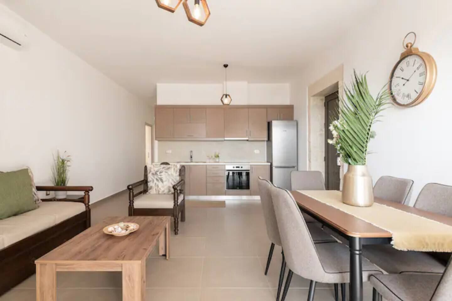 A modern living room with a sofa, dining table, and chairs. There is a kitchen in the background with cabinets and appliances.