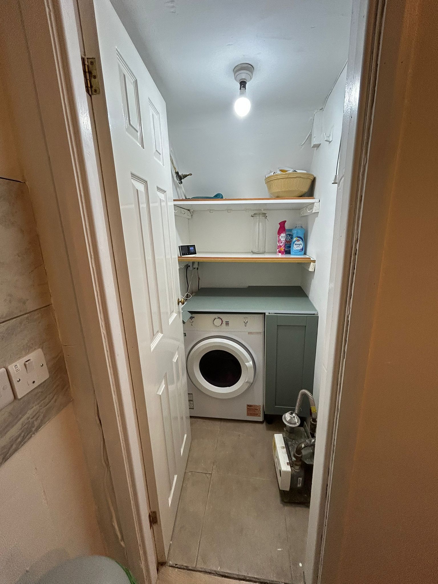 A compact laundry room with a washing machine, shelves with toiletries, and a ceiling light.