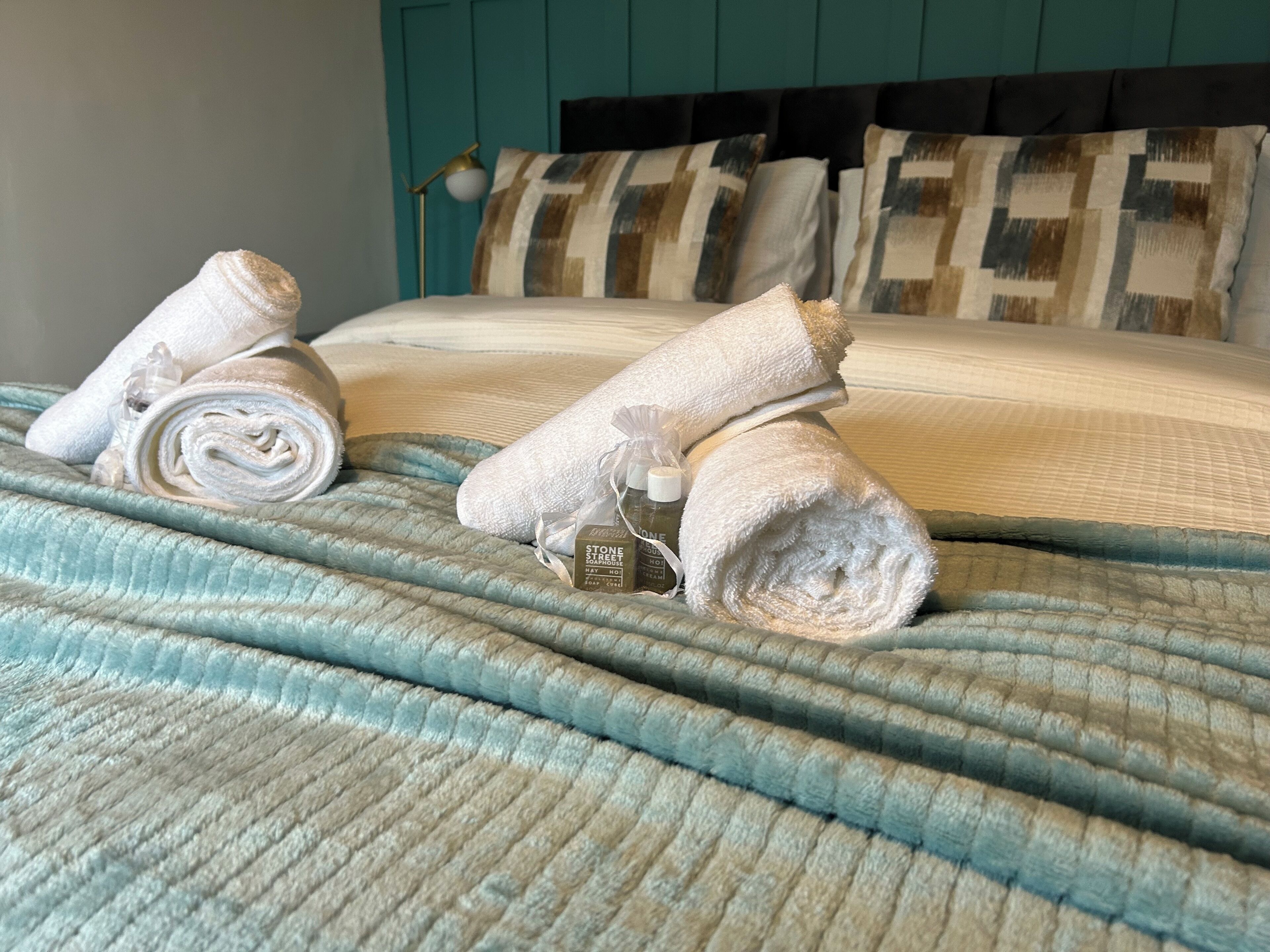 Una cama bien hecha con toallas enrolladas y una botella de loción, frente a una pared color verde azulado y un cabecero estampado.