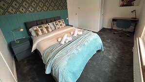 A neatly made bed with a patterned headboard, a desk with a chair, and a wall-mounted artwork.
