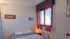 A small, simple room with a bed, a nightstand, a window with curtains, and a wall-mounted light. - Seignosse Penon - T2 Atlantic Coast Near Beach (Seignosse)