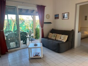 A living room with a sofa, a small table, a sliding glass door leading to an outdoor area with chairs and a table, and a bed in the adjacent room. - Villa 6 pers Aircon - Direct Beach Access - Pool (Gruissan)