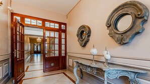 Um corredor amplo com porta de madeira, um console de mármore com objetos decorativos e dois espelhos de parede elaborados.