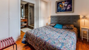 Quarto com uma cama grande, mesa de cabeceira, um banco, guarda-roupa e área de chuveiro.