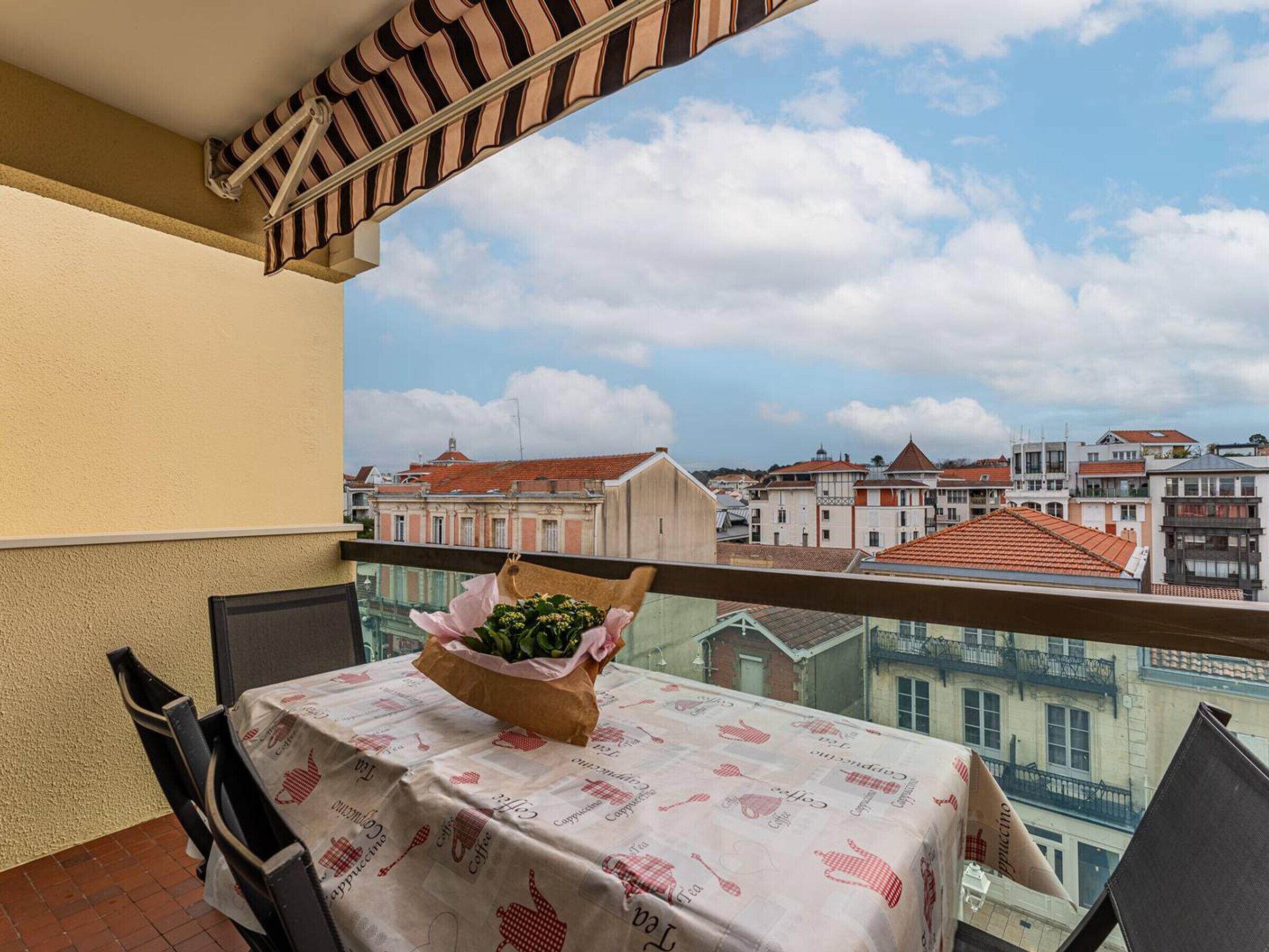 A balcony with a table covered in a tablecloth with a coffee theme, a chair, and a bouquet of flowers.