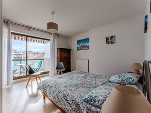 A bedroom with a bed, a chair, a table, and a view of the city through large windows. - Spacious T3 apartment with parking, balconies, 20m from Arcachon beach (Arcachon)