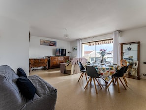 A spacious living area with a grey sofa, wooden dining table, and chairs. A TV is mounted on the wall, and there is a window with a view of buildings. - Spacious T3 apartment with parking, balconies, 20m from Arcachon beach (Arcachon)