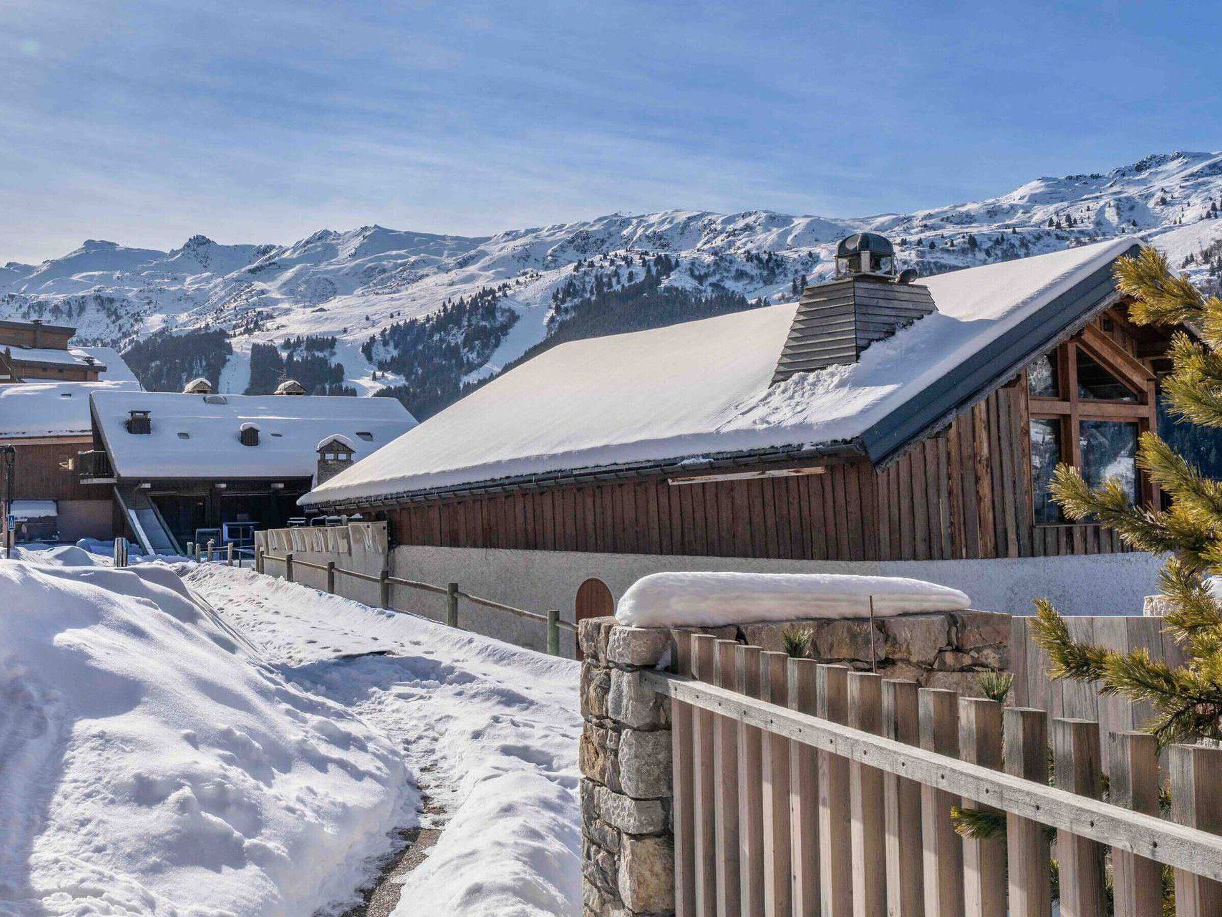 A wooden cabin with a snow-covered roof, surrounded by a stone wall and wooden fence, set against a backdrop of snow-covered mountains.