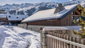 A wooden cabin with a snow-covered roof, surrounded by a stone wall and wooden fence, set against a backdrop of snow-covered mountains. - Rental in Méribel - Altiport Address (Méribel)