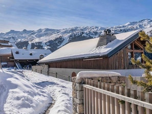 Eine Holzhütte mit schneebedecktem Dach, umgeben von einer Steinmauer und einem Holzzaun, vor einer Kulisse verschneiter Berge.