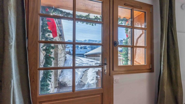 A wooden door with a glass panel, leading to a snowy outdoor area with trees and a mountain view. - Rental in Méribel - Altiport Address (Méribel)