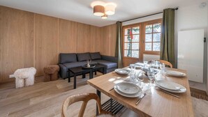 A modern living room with a wooden floor, a sofa, a dining table set for four, and a window with curtains. - Rental in Méribel - Altiport Address (Méribel)