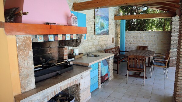 A rustic outdoor kitchen with a brick oven, wooden furniture, and a stone wall backdrop.