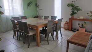 Et spiseområde med et stort træbord omgivet af fletstole, et lille sidebord med en plante og et vindue med lette gardiner.