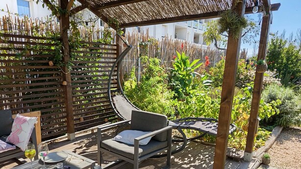 En træterrasse med et pergolaoverdækning, et polstret udendørs opholdsområde og en have med forskellige planter.