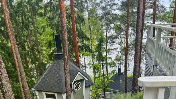 Una terraza de madera que lleva a una pequeña cabaña con chimenea, rodeada de árboles altos y una masa de agua.