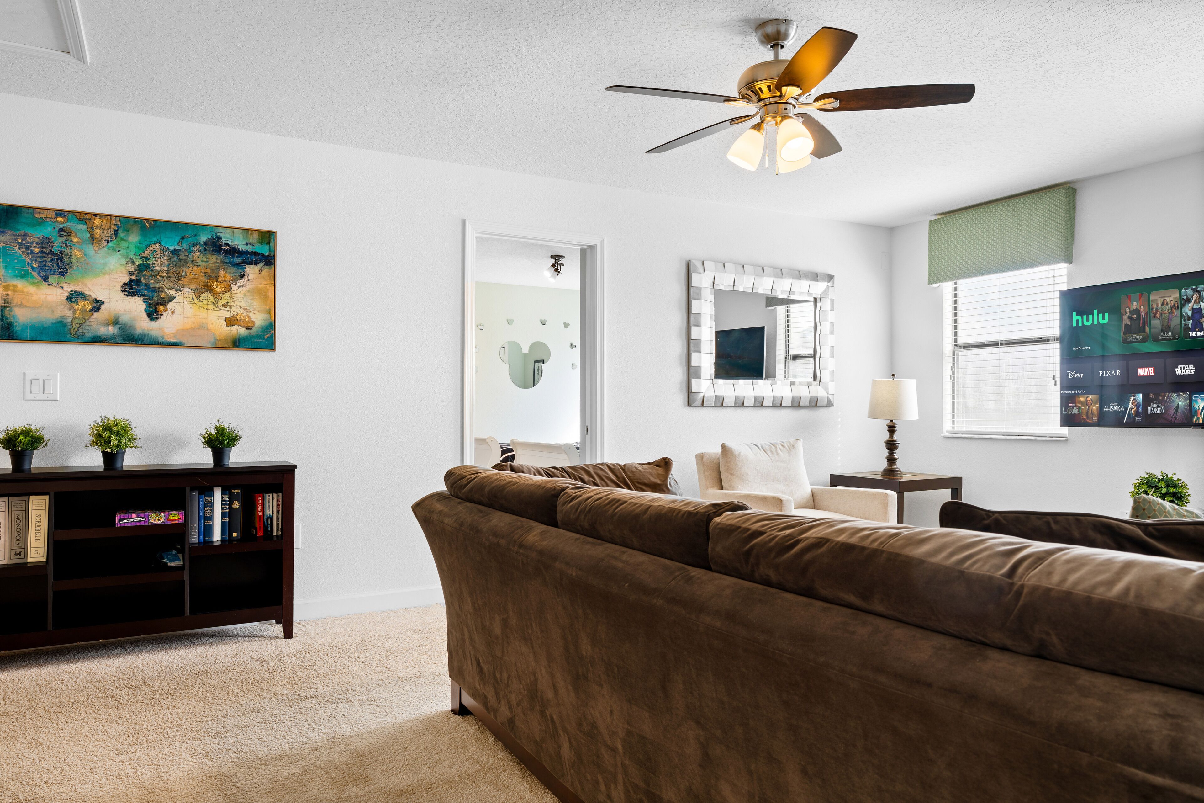 A living room with a brown sofa, a ceiling fan, a world map painting, a bookshelf, and a TV displaying the Hulu app.