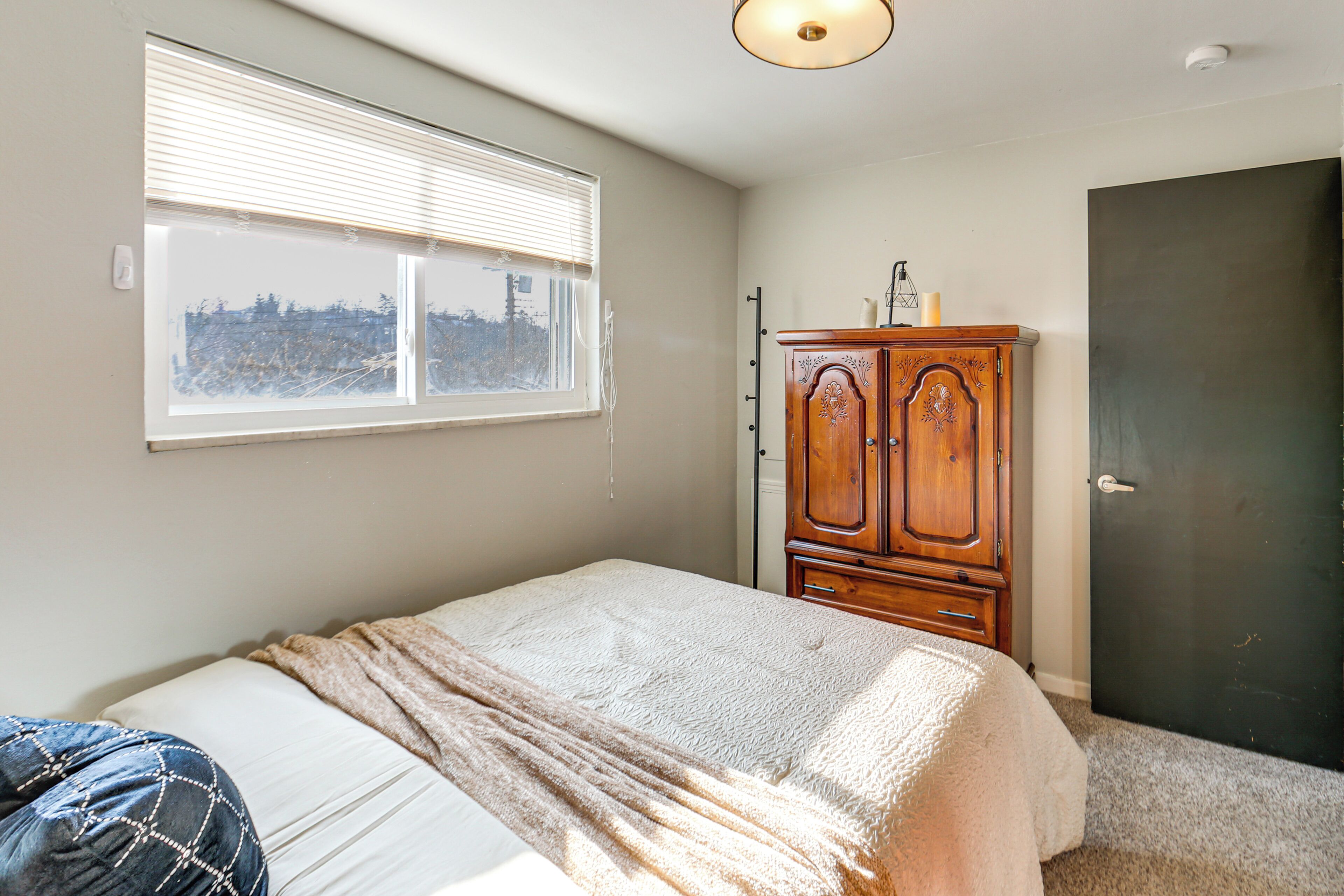 A bedroom with a bed, a window with blinds, a wooden wardrobe, and a door.