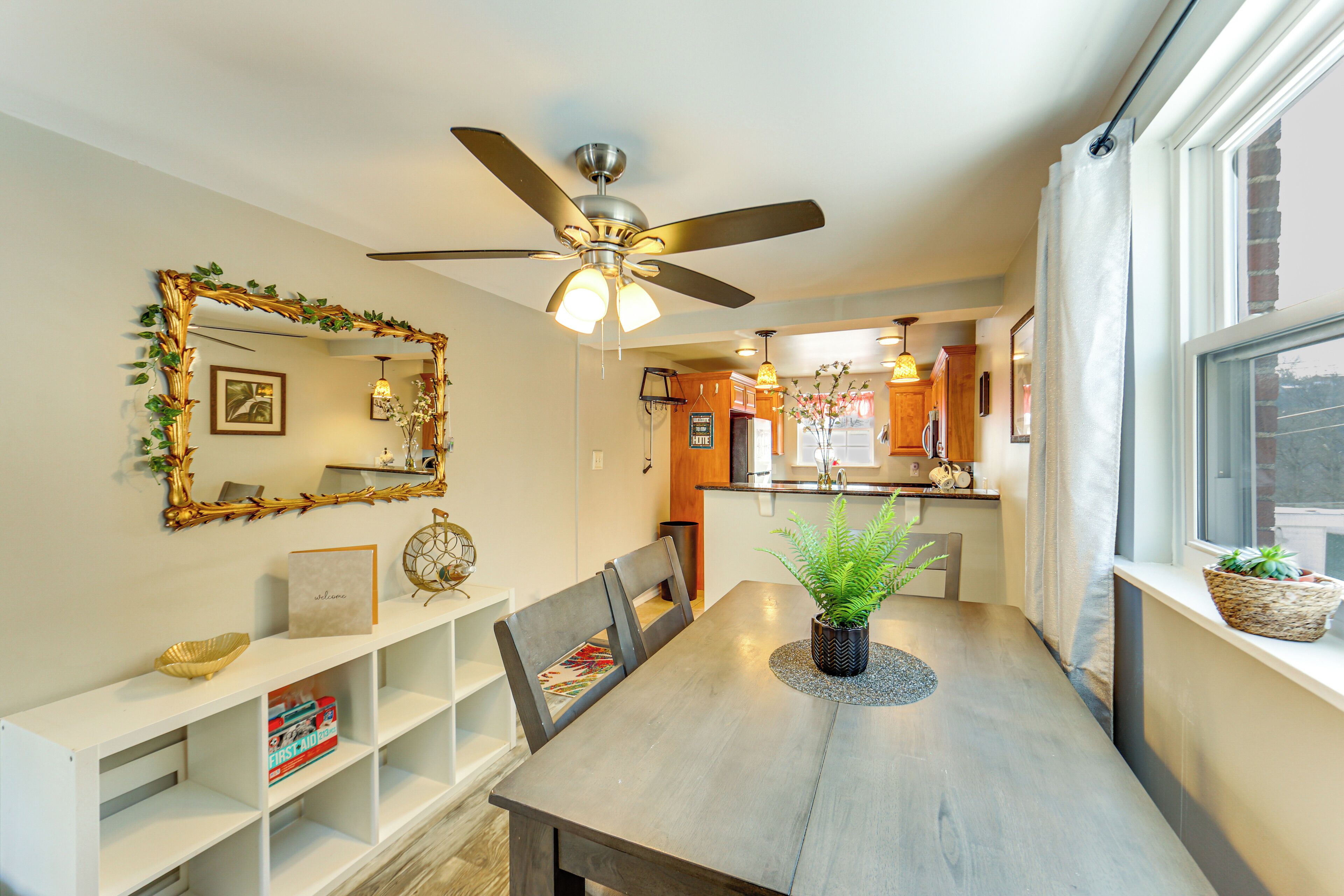 A dining area with a wooden table, chairs, a ceiling fan, a mirror with a decorative frame, and a potted plant.
