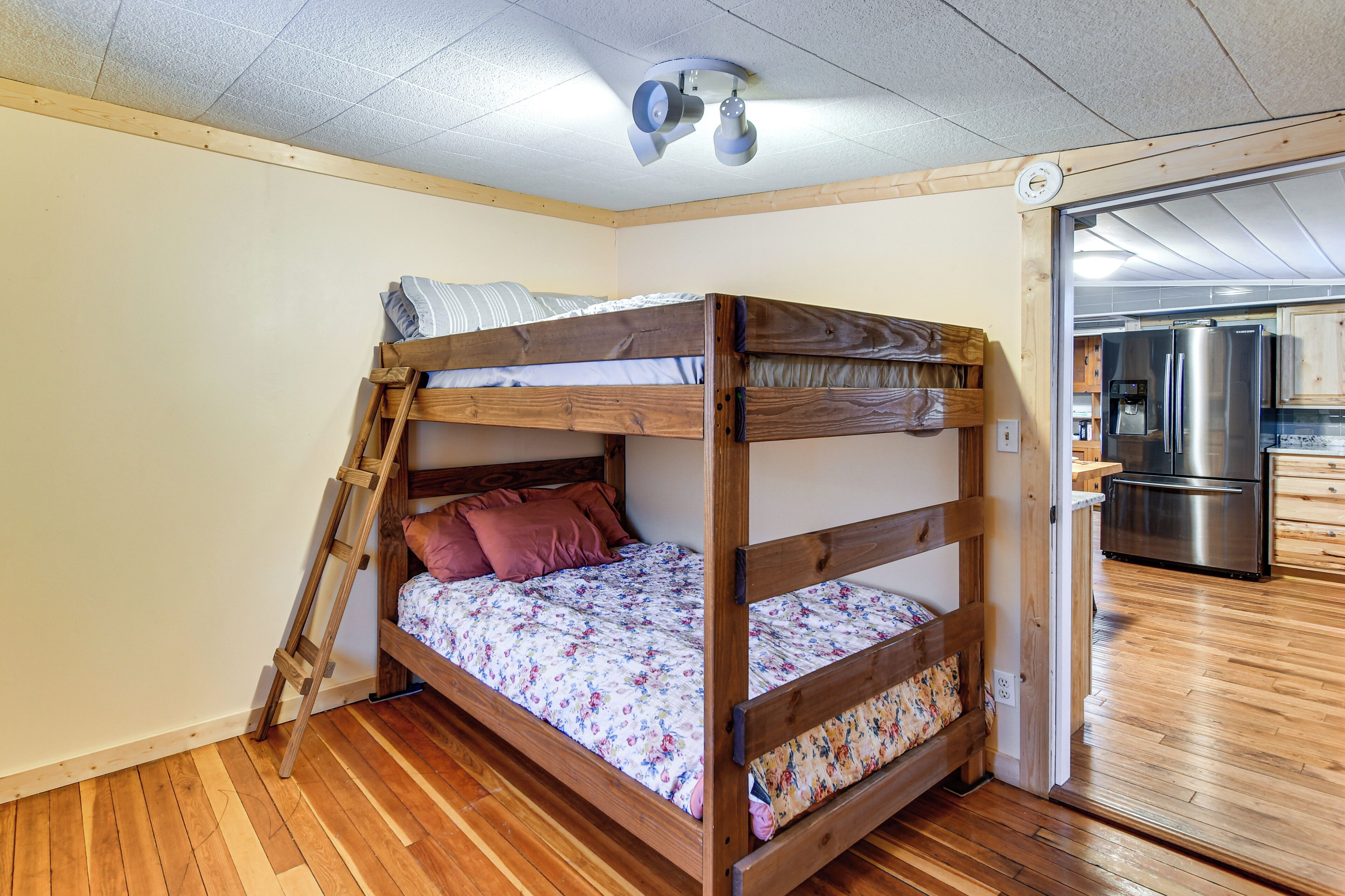 A bunk bed with a ladder, wooden flooring, and a kitchen visible through an open door.