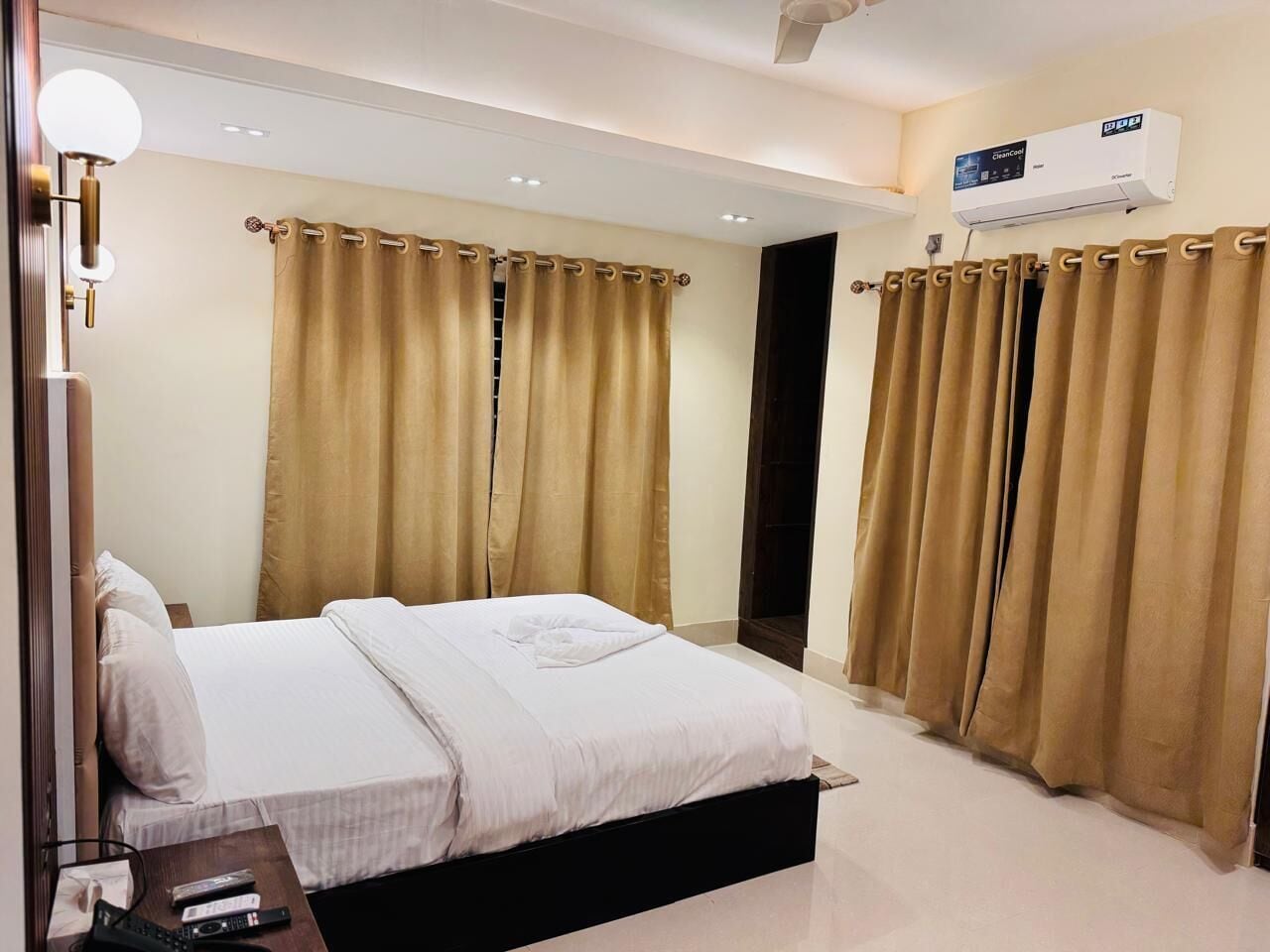 A hotel room with a bed, curtains, a bedside table, a lamp, and an air conditioning unit.