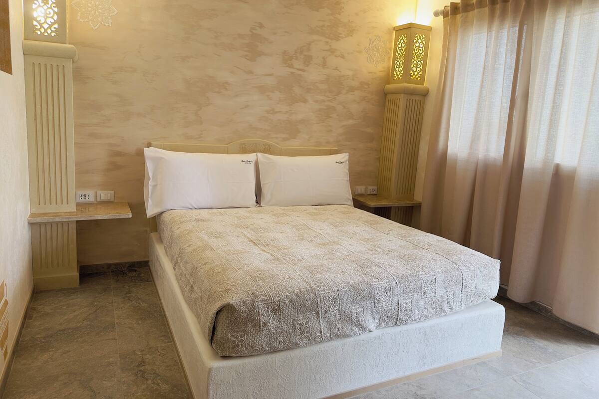 A neatly arranged bedroom with a large bed, two bedside tables, a wall-mounted light fixture, and a window with curtains.