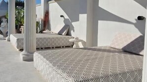 Un espacio al aire libre con asientos iluminados por el sol, columnas blancas y cojines estampados.
