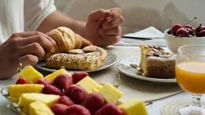 Un desayuno con cruasanes, fruta, una porción de pastel y un vaso de jugo de naranja sobre una mesa junto a una ventana.