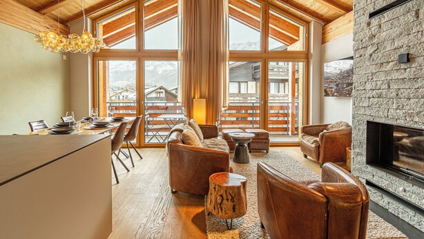 Un vaste espace de vie doté d’un haut plafond en bois, de grandes fenêtres, d’une cheminée et d’un coin repas avec une table dressée pour quatre personnes.