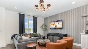 A modern living room with a grey sofa, brown leather armchair, and a coffee table. A flat-screen TV mounted on the wall displays a basketball game. - 3 Side-by-Side Homes | 12BR | 3 Rooftop Hot Tubs (nashville)