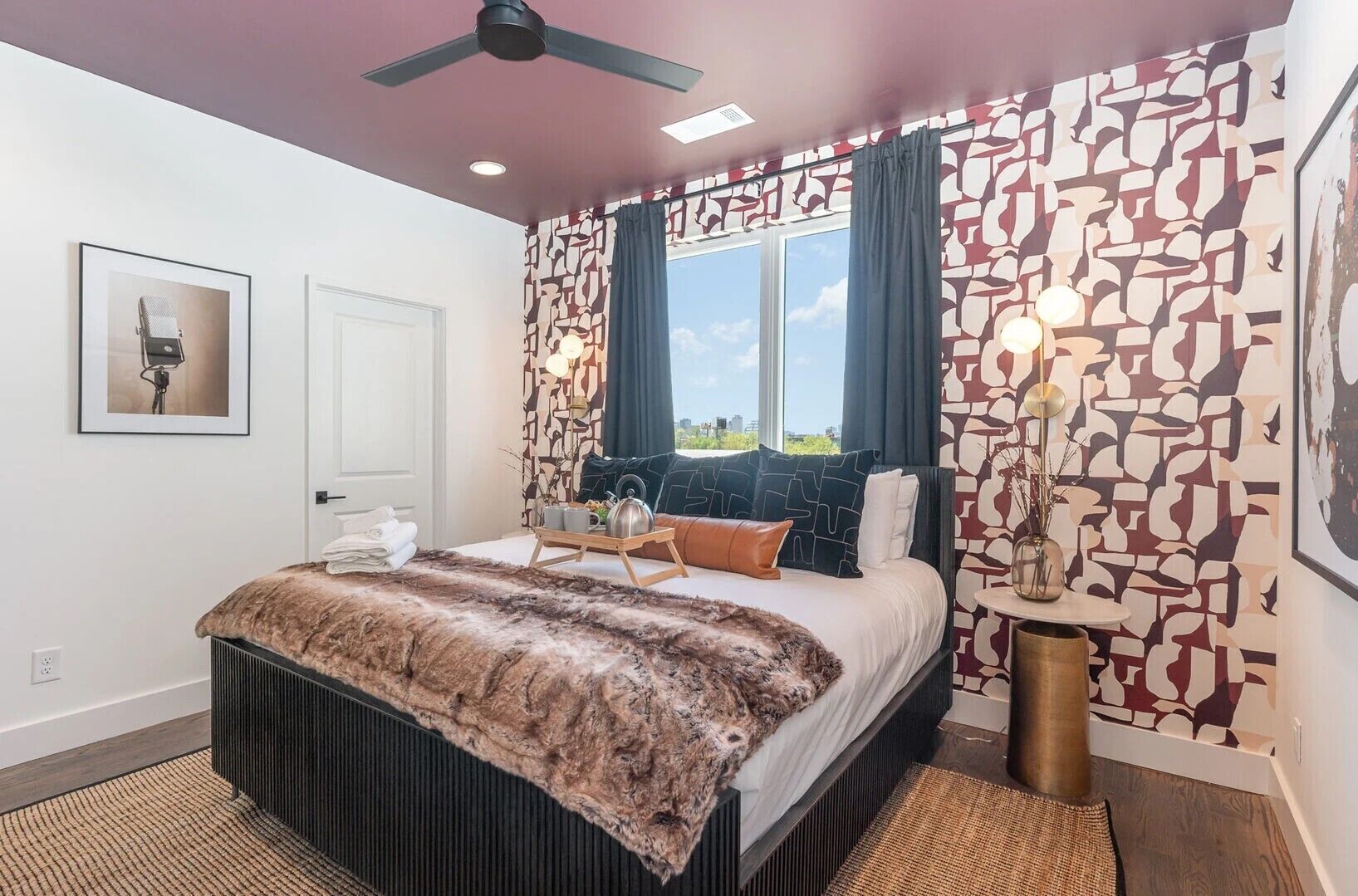 A bedroom with a bed, a ceiling fan, a window with curtains, a wall with a patterned design, and a framed picture on the wall.