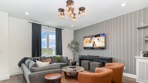 Een moderne woonkamer met een grijze bank, een fauteuil van bruin leer en een salontafel. Een flatscreen televisie aan de muur toont een basketbalwedstrijd.