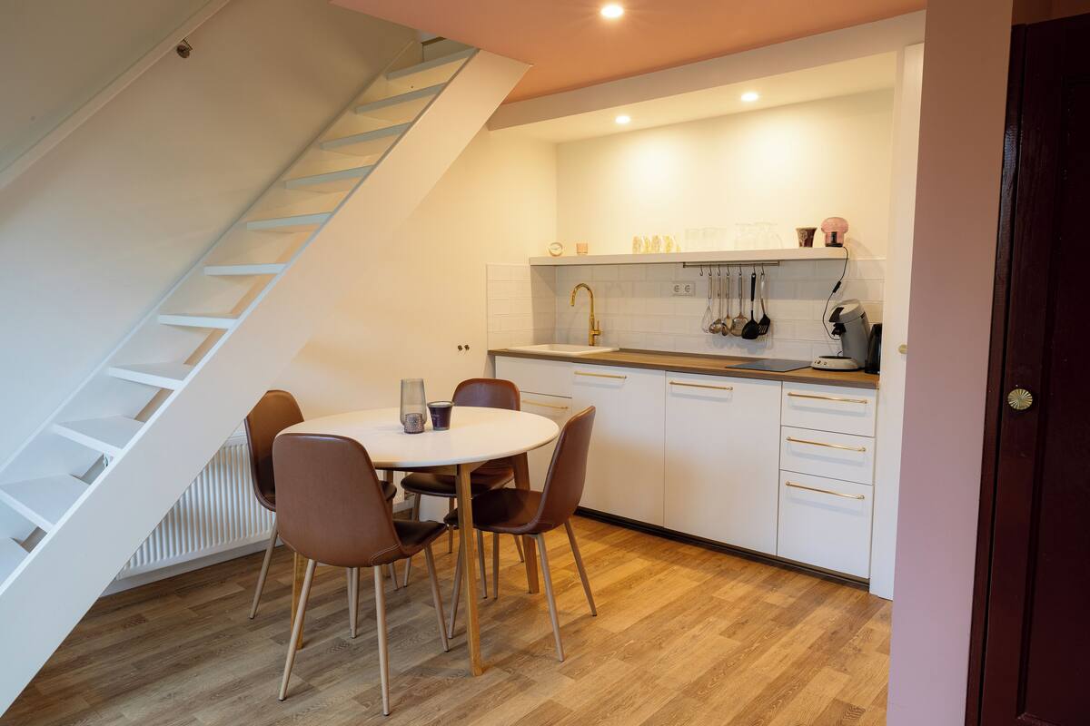 A modern kitchen with white cabinets, a white countertop, and a built-in oven. There is a dining table with chairs and a staircase leading to an upper level.