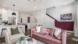Ett modernt vardagsrum med en rosa soffa, en vit fåtölj och ett runt soffbord. I bakgrunden finns ett kök med vita skåp och vitvaror i rostfritt stål.