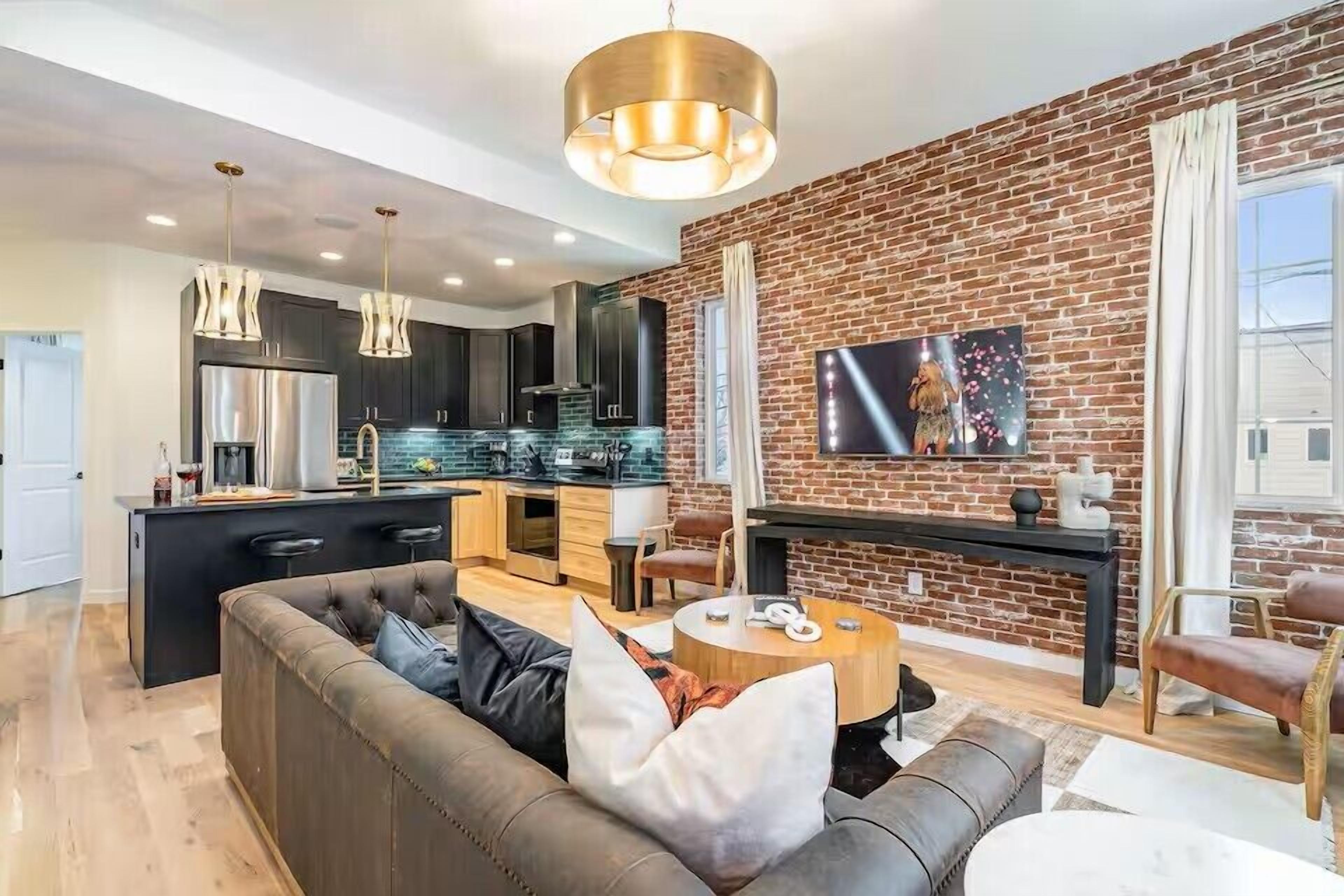 A modern kitchen with a brick wall, a dining area with a round table, and a living room with a sofa.