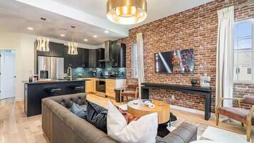A modern kitchen with a brick wall, a dining area with a round table, and a living room with a sofa.