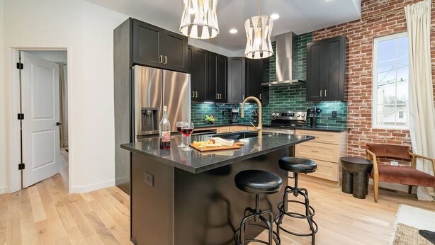Una cocina moderna con armarios oscuros, una isla central con taburetes, electrodomésticos de acero inoxidable y una pared decorada con ladrillo.