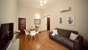 Un salon avec un canapé marron, des chaises blanches, une table à manger et un téléviseur. Des tableaux encadrés ornent le mur et un climatiseur est installé au-dessus du canapé.