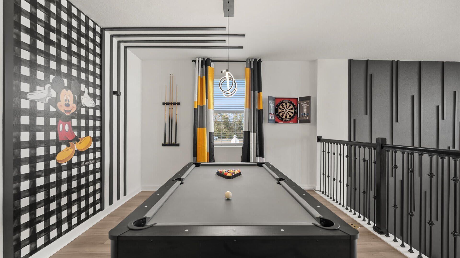 A pool table in a room with a checkered wall, a dartboard, and a window with curtains.