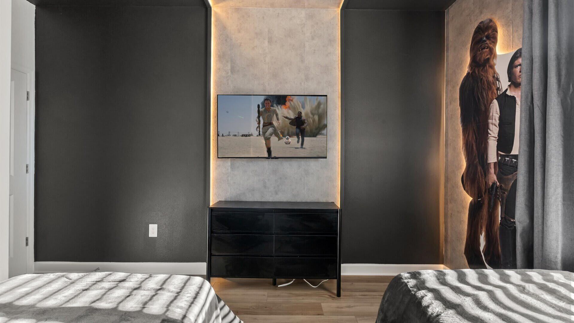 Een moderne slaapkamer met een groot flatscreen-televisie aan de muur, een zwarte tv-meubel eronder en een Star Wars-muurdecoratie aan de rechtermuur.
