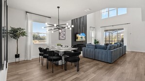 A modern living room with a grey sofa, a dining area with a table set for four, and a large window with a view of the outdoors. - Family 5BR Villa w Pool Theatre Reunion Village (Kissimmee)