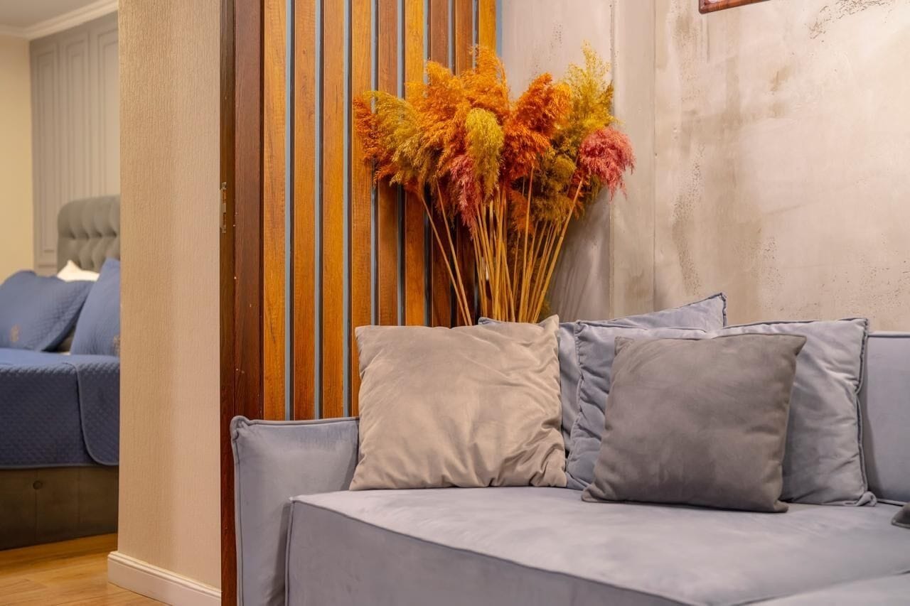 Un salon avec un canapé gris, un mur recouvert de lambris en bois et un vase de fleurs séchées.