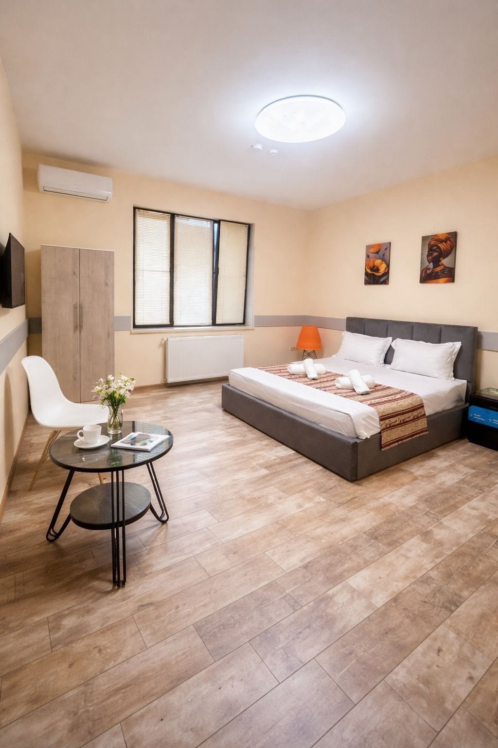 A hotel room with a bed, a small table with a cup and flowers, a chair, a radiator, and a window with blinds.
