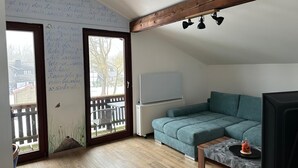 Una habitación acogedora con un sofá color turquesa, una mesita pequeña y un televisor. Hay una ventana con vistas al exterior, una pizarra con texto y una viga de techo de madera.