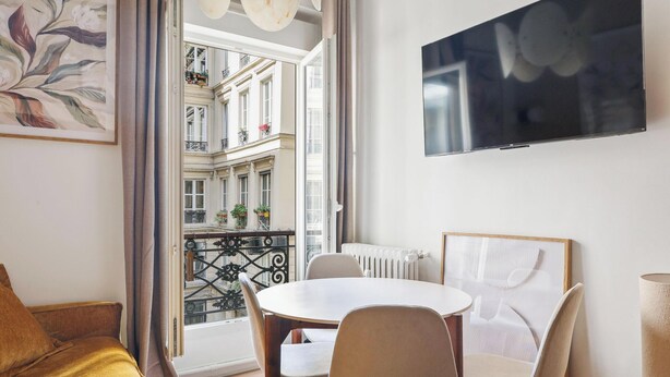 Ein Zimmer mit Esstisch, Stühlen, Fernseher, Heizkörper und einem Balkon mit Pflanzen.