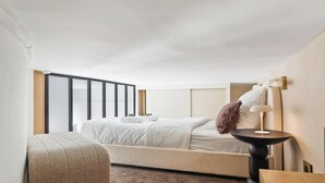 Ein Hotelzimmer mit Bett, Sessel, Nachttisch und einem großen Fenster mit Jalousien.