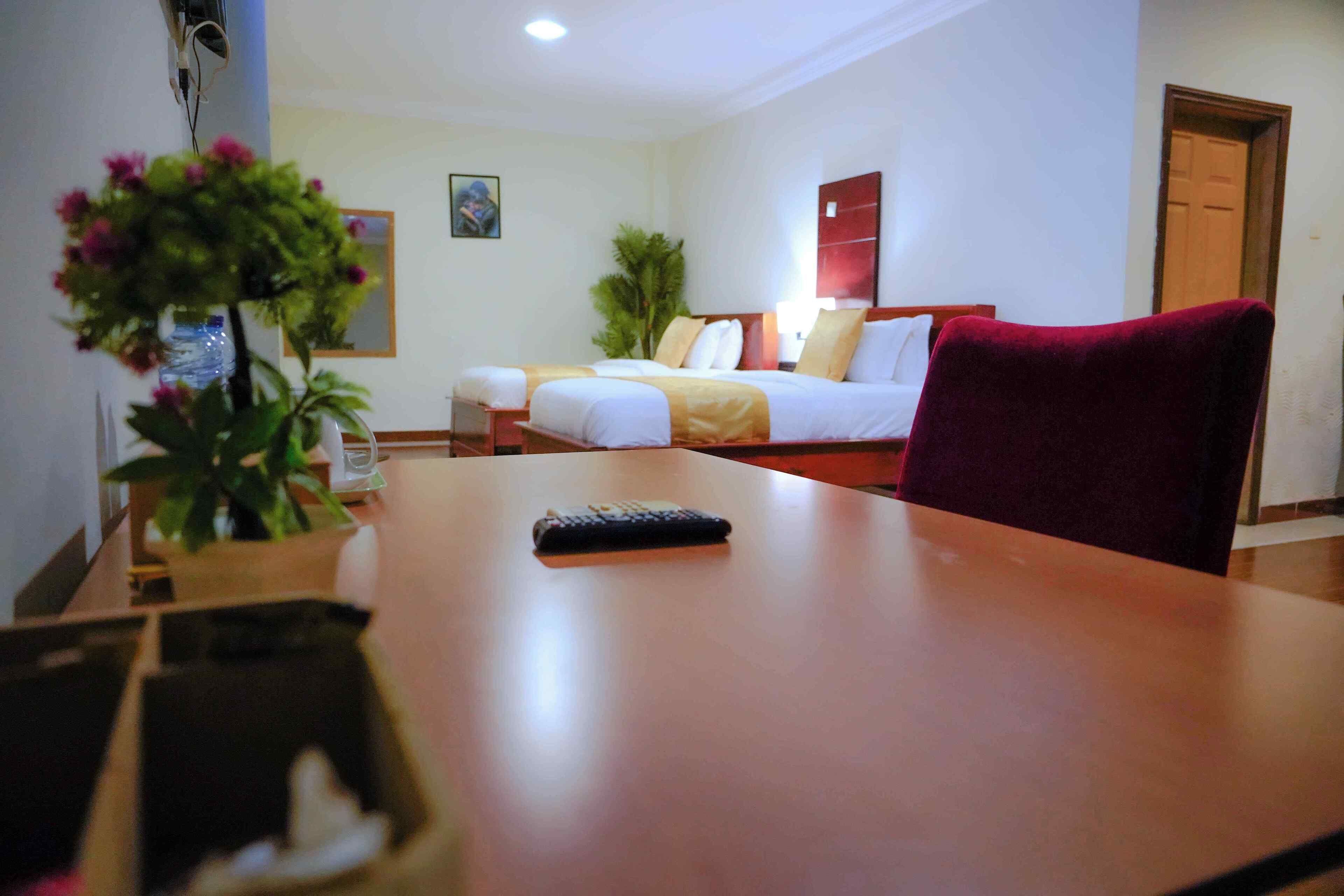 A hotel room with a wooden table, a bed with white bedding, a red chair, and a potted plant.