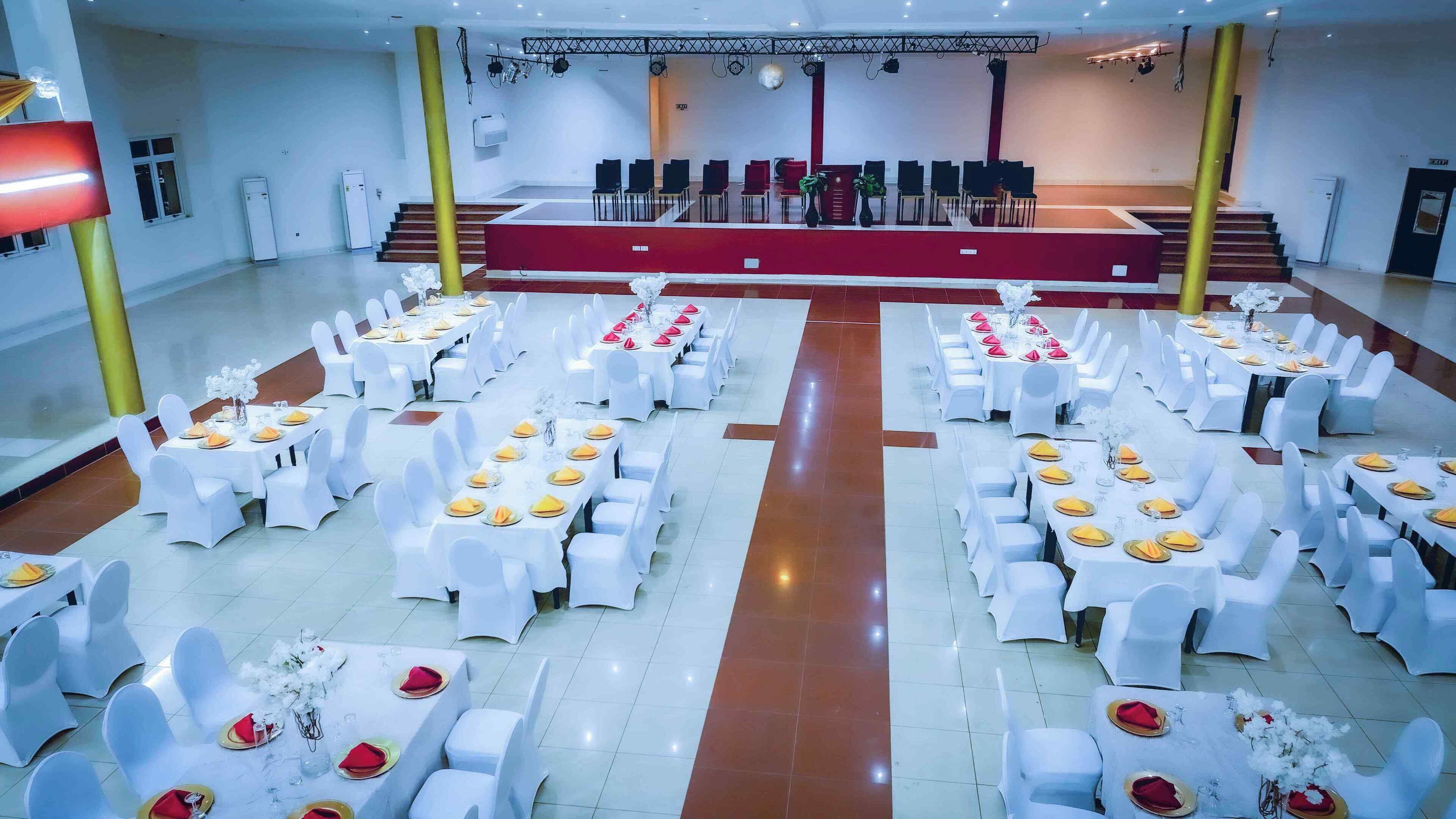 Une salle de banquet dotée de nombreuses tables rondes, chacune recouverte d’une nappe blanche, agrémentée de chaises blanches et de serviettes jaunes.